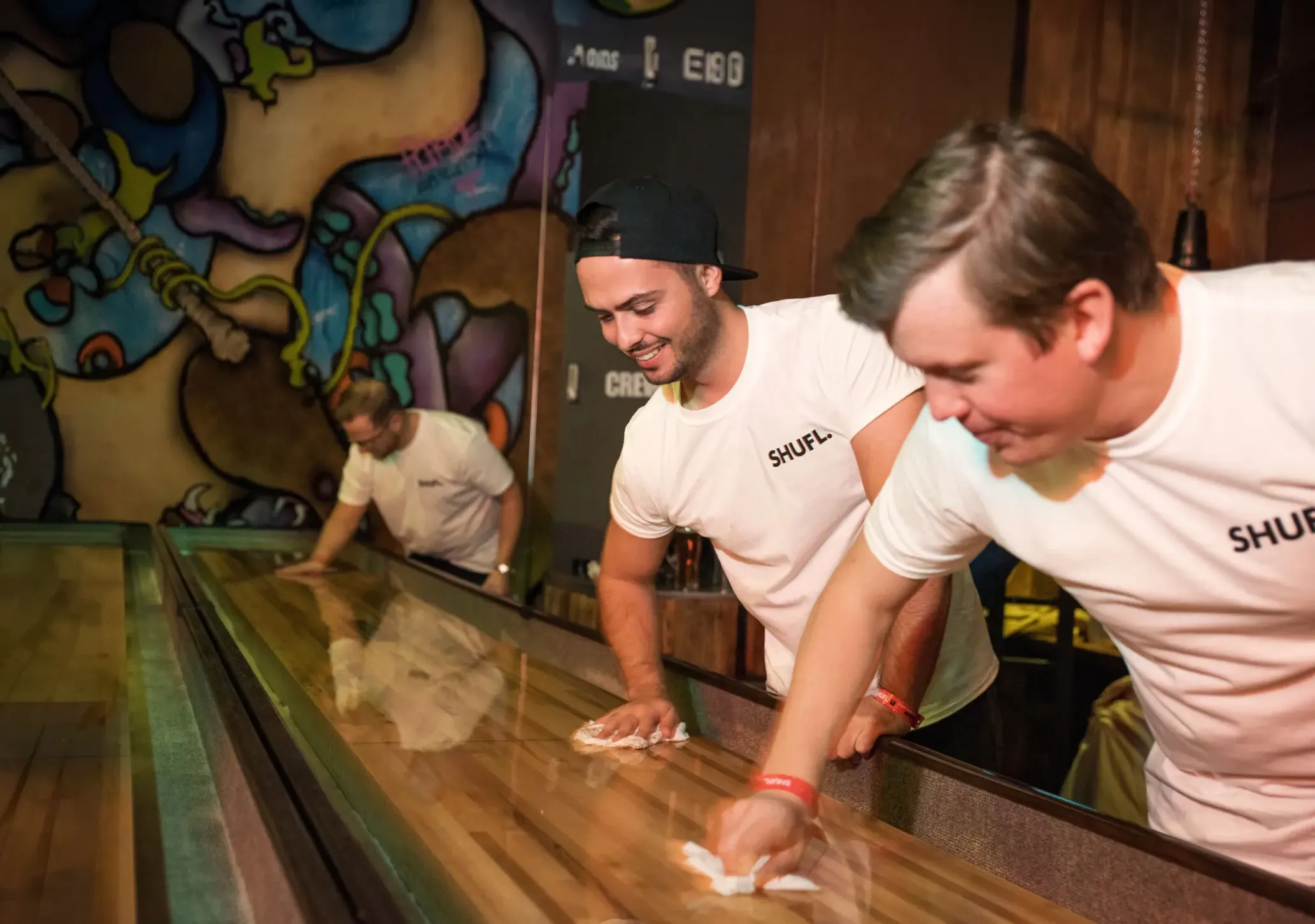 Two venue staff wiping down a SHUFL shuffleboard table in a graffiti-walled bar